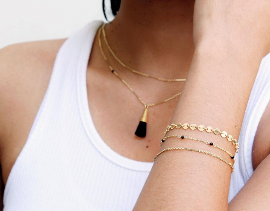 Close up of woman wearing sustainably and ethically made jewelry from wearwell personal stylist subscription membership. She wears two gold chain necklaces one with black tassel pendant, two gold chain bracelets and one gold bracelet with black beads.