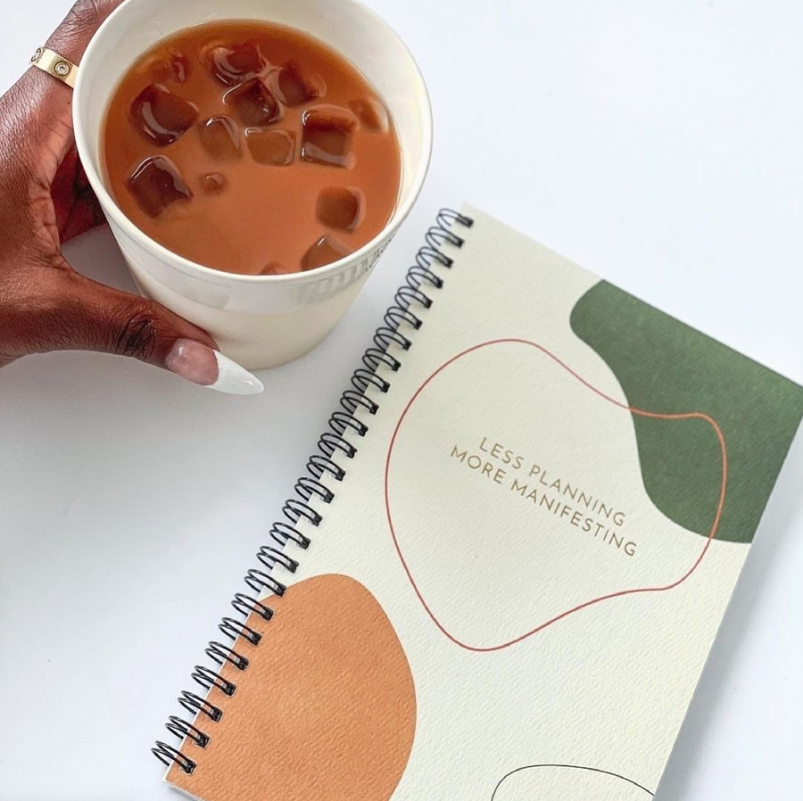 Close up of spiral bound notebook with green, orange and pink abstract cover design made from responsibly sourced recycled paper produced by black owned brand and a woman reaching for beverage.
