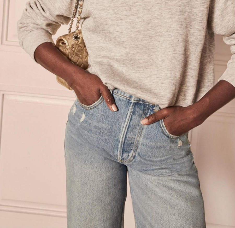 Close up of woman wearing distressed light wash jeans, long sleeve oatmeal sweater and gold purse. All clothing and accessories from wearwell personal stylist subscription membership and made in ethical and fair trade facilities.