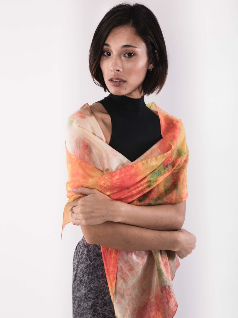 Woman wearing a black mock neck top, dark grey skirt and a hand dyed orange, green, and yellow scarf around her shoulders, all ethically made in a fair trade facility and from wearwell personal stylist subscription membership.
