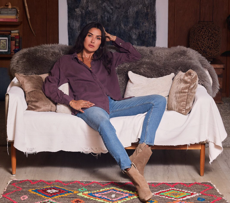 Woman siting on couch wearing sustainable ethical clothing and accessories from wearwell personal stylist subscription membership including a purple button down blouse, blue skinny jeans and brown boots made at a fair trade facility.