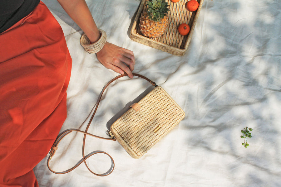 Woman wearing red skirt, black top and woven bracelet sitting on blanket next to woven rattan and leather purse and rattan tray made by skilled artisans at fair trade and ethical facility. All are from wearwell personal stylist membership.