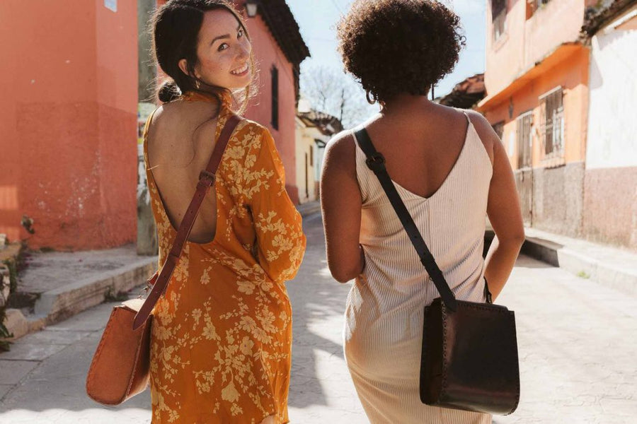 Two women walking, one wearing a sustainably made yellow floral dress, the other in a white ethically produced slip dress, both wearing hoop earrings and carrying artisan made leather crossbody bags from wearwell personal stylist subscription membership.