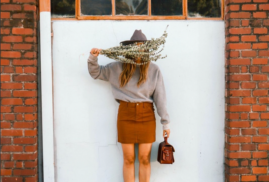 Woman standing in front of an exterior wall wearing a grey sweatshirt, grey brimmed hat and earth tone brown organic cotton skirt, holding a small leather purse. All sustainable ethical clothing and accessories from wearwell personal stylist subscription.